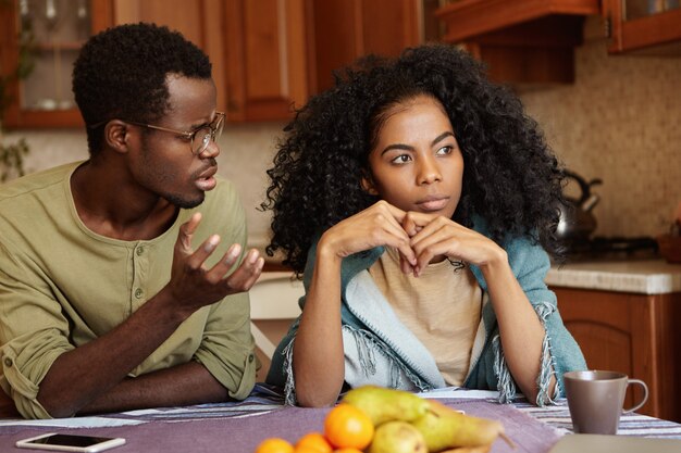 ¿Por qué me hiciste esto? Indignado, deprimido, joven afroamericano con gafas tratando de conversar con su esposa indiferente que lo engañó. Problemas de relación e infidelidad