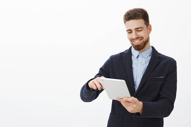 Qué placer mirar la cuenta bancaria llena de dinero. Encantado de guapo y exitoso hombre de negocios con barba y peinado prolijo en traje sosteniendo tableta digital sonriendo complacido mirando la pantalla del dispositivo