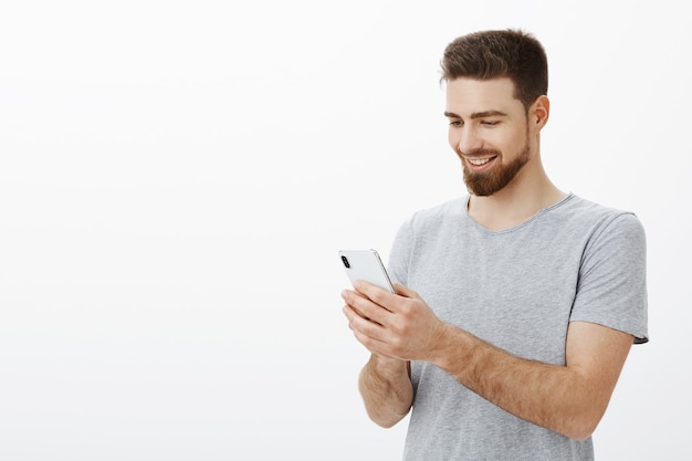 Qué placer mirar la cuenta bancaria llena de dinero. Retrato de feliz elegante y guapo modelo masculino con barba sosteniendo smarpthone mirando la pantalla del teléfono móvil con alegría y sonrisa alegre