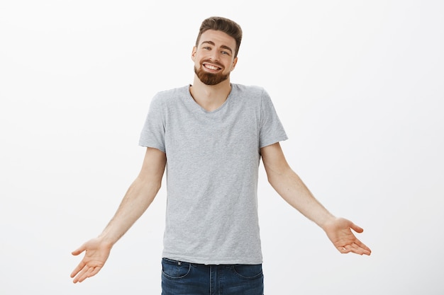 Foto gratuita ¿qué puedo decir? soy el novio perfecto. carismático y encantador chico europeo con barba encogiéndose de hombros con los brazos abiertos con una linda sonrisa encantadora recibiendo cumplidos por su buen trabajo sintiéndose seguro de sí mismo