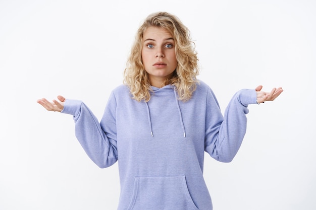 Que sé yo. Retrato de mujer rubia europea linda confundida e inconsciente con ojos azules de pie interrogada con mirada despistada encogiéndose de hombros y extendiendo las manos hacia los lados con consternación incierta sobre la pared blanca