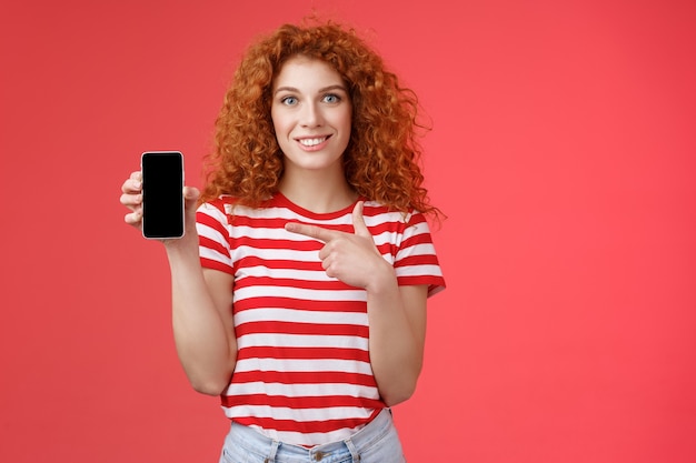 ¿Qué tal esta aplicación? Pelirroja rizada amigable linda chica ayuda a un amigo a encontrar tienda en línea oufit sostenga la pantalla del teléfono del teléfono inteligente señalando sonriendo ampliamente recomendar filtro de fotos de redes sociales, fondo rojo.