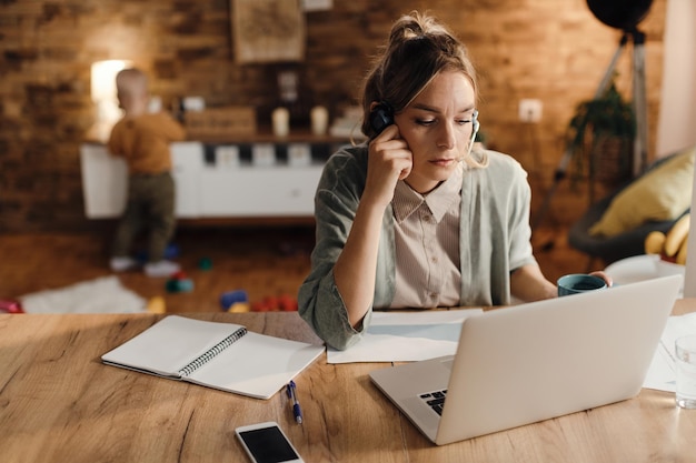 Quédese en casa madre trabajando en una computadora mientras su hijo juega en el fondo