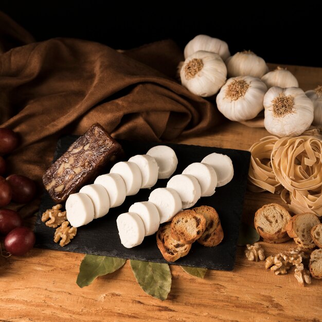 Queso de cabra fresco en bandeja de pizarra con rebanadas de pan y nueces cerca de pastas crudas y bulbos de ajo