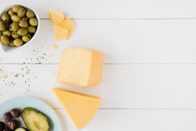 Foto gratuita queso cheddar con aceitunas verdes y rebanada de aguacate en mesa de madera blanca