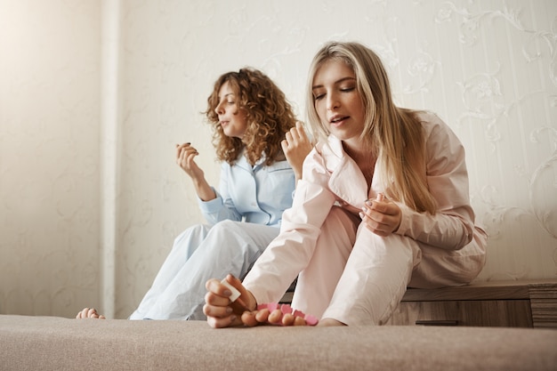 ¿Quién necesita salones de belleza cuando puedes hacer uno en casa? Retrato de dos mejores amigos sentados en la habitación y pintando las uñas de los pies, vistiendo ropa de dormir y sintiéndose cómodos, teniendo una fiesta femenina