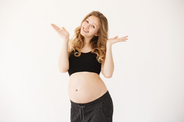 Quién sabe. Hermosa rubia alegre mujer embarazada europea en traje cómodo con el vientre abierto extendiendo las manos con expresión alegre.