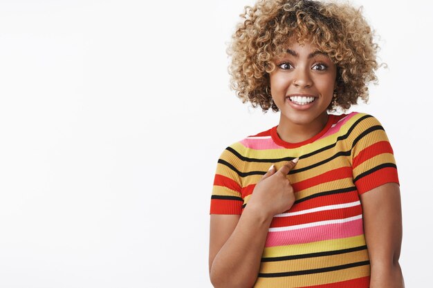 Quien yo. Retrato de niña afroamericana encantadora, linda y despreocupada sorprendida señalando cuestionada y emocionada de sí misma como mencionada o elegida sonriendo ampliamente posando sobre una pared blanca