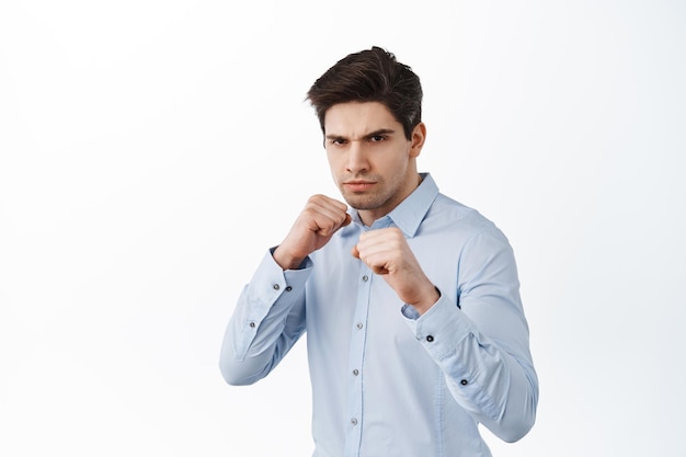 Quieres pelear. Trabajador de oficina serio en camisa levantando los puños, de pie en pose de boxeador defensivo, boxeo de sombra, de pie sobre fondo blanco