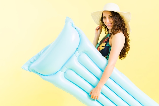 ¿Quieres venir a la piscina? Mujer caucásica coqueta que lleva un colchón inflable. Mujer joven con un sombrero de verano caminando a la piscina