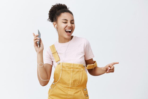 Quiero bailar con alguien. Retrato de mujer afroamericana atractiva feliz alegre en peto con estilo amarillo, moviéndose al ritmo de la música, escuchando canciones en auriculares, sosteniendo el teléfono celular