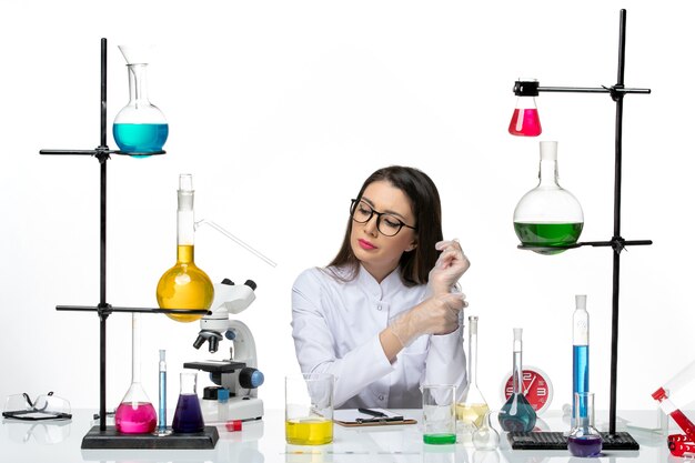 Química de vista frontal en traje médico blanco sentado con diferentes soluciones sobre fondo blanco laboratorio covid de virus pandémico de ciencia