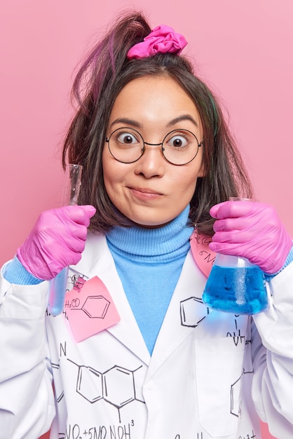 químico examina líquido químico en matraces realiza experimento en laboratorio usa anteojos redondos bata blanca guantes de goma aislados en pared rosa