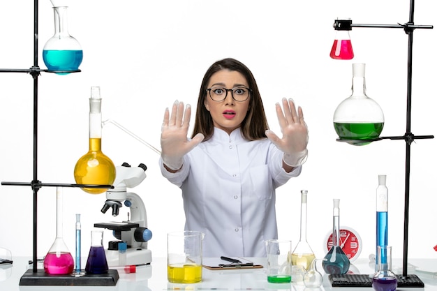 Químico femenino de vista frontal en traje médico blanco sentado con soluciones en el laboratorio de covid de virus pandémico de ciencia de escritorio blanco