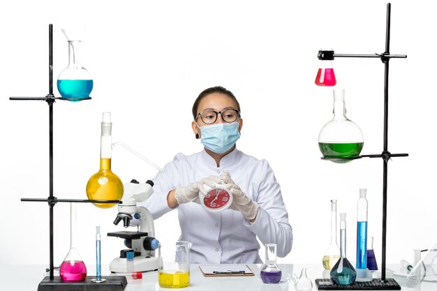 Químico femenino de vista frontal en traje médico con máscara sosteniendo relojes en el laboratorio de virus de escritorio blanco química covid splash