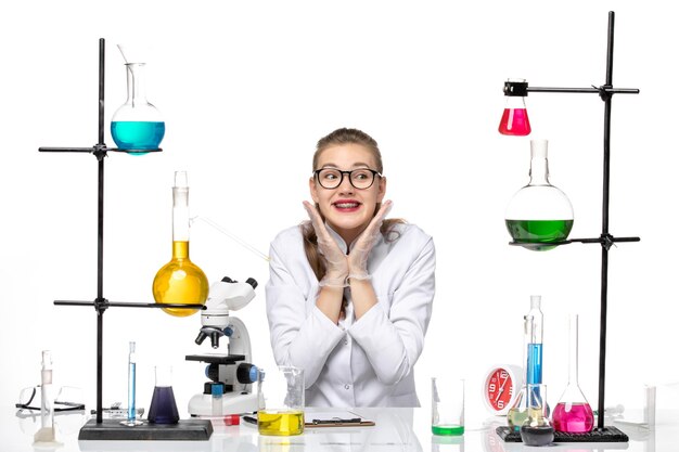 Químico femenino de vista frontal en traje médico sentado con soluciones y sonriendo sobre fondo blanco química pandemia covid-virus