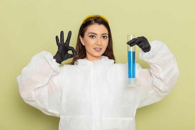 Químico femenino de vista frontal en traje de protección especial con matraz con solución azul sonriendo sobre la superficie verde