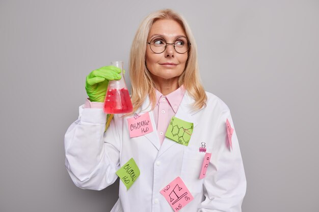 El químico lleva gafas redondas bata blanca con pegatinas pegadas fórmulas escritas sostiene frasco de vidrio con líquido rojo después de mezclar reactivos aislados en gris