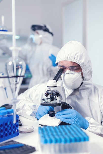 Químico con mascarilla y desarrollo de vacunas ppe para covid mirando a través del microscopio. Científico en traje de protección sentado en el lugar de trabajo utilizando tecnología médica moderna durante la epidemia global.