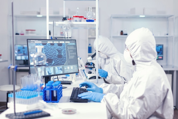 Químico en traje de ppe escribiendo en el teclado comprobando el desarrollo de virus en el laboratorio equipado. Ingeniero médico que usa una computadora durante una pandemia global con coronavirus vestido con un mono.