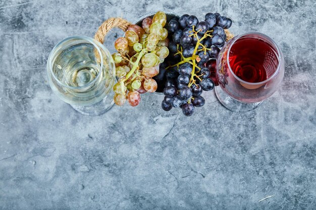 Racimo de uvas blancas y negras y dos vasos de vino blanco y rojo sobre azul.