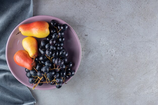 Racimo de uvas con peras rojo-amarillo en placa violeta sobre mesa de piedra.