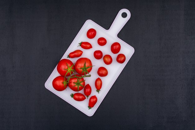 Racimos de tomate y tomate cherry rojo sobre tabla de cortar blanca sobre superficie de piedra negra