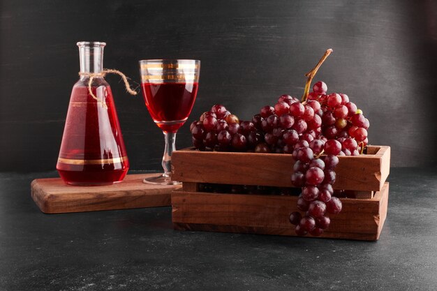 Racimos de uva roja en una bandeja de madera con una copa de vino sobre negro.