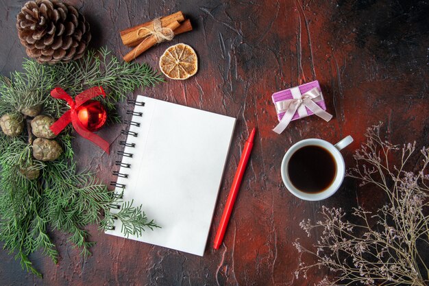 Ramas de abeto y cuaderno de espiral cerrado con bolígrafo, canela, limas, cono de coníferas y una taza de té negro sobre fondo oscuro