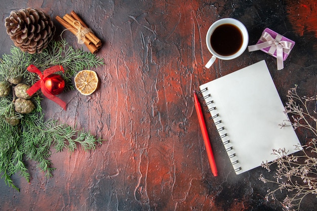 Ramas de abeto y cuaderno de espiral cerrado con bolígrafo, canela, limas, cono de coníferas y una taza de té negro sobre fondo oscuro