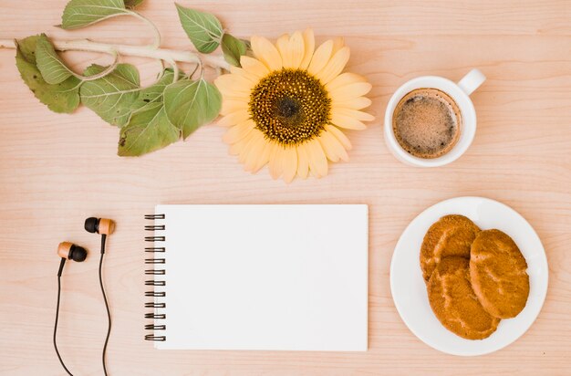 Ramita de girasol; taza de café; auricular; Galletas y bloc de notas de espiral en blanco en el escritorio de madera