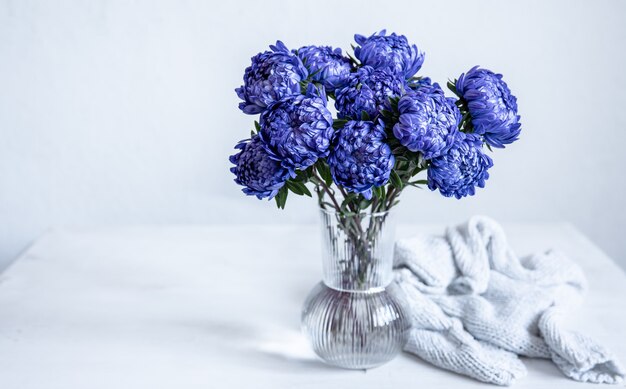Un ramo de crisantemos azules en un jarrón de vidrio y un elemento tejido sobre un fondo blanco, copie el espacio.