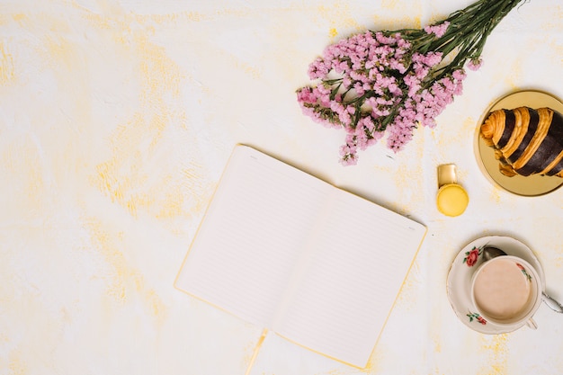 Ramo de flores con cuaderno, café y croissant en mesa.