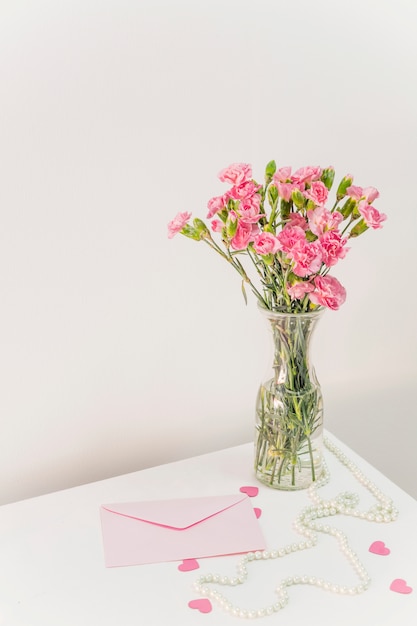 Ramo de flores en florero cerca de sobre, corazones de papel y cuentas en la mesa