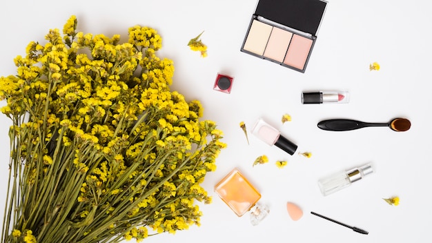 Ramo de flores de limonio amarillo; botella de esmalte de uñas; frascos de perfume; Lápiz labial y pincel de maquillaje y polvos faciales compactos sobre fondo blanco