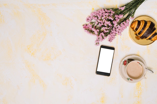 Ramo de flores con smartphone, café y croissant en mesa.