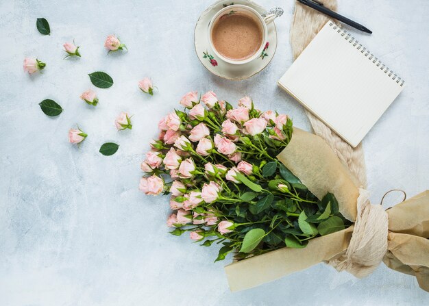 Ramo fresco de rosas rosadas con la taza de café; Bloc de notas de espiral y pluma sobre fondo con textura