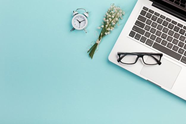 Ramo de lirio de los valles con reloj despertador, lentes y computadora portátil en el escritorio de la oficina azul