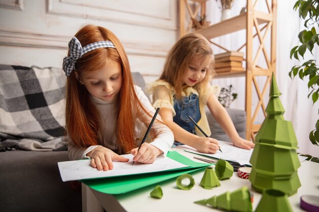 Árbol de Navidad. Dos niños pequeños, niñas juntas en creatividad. Los niños felices hacen juguetes hechos a mano para juegos o celebración de Año Nuevo. Pequeños modelos caucásicos. Infancia feliz, preparación para la celebración.