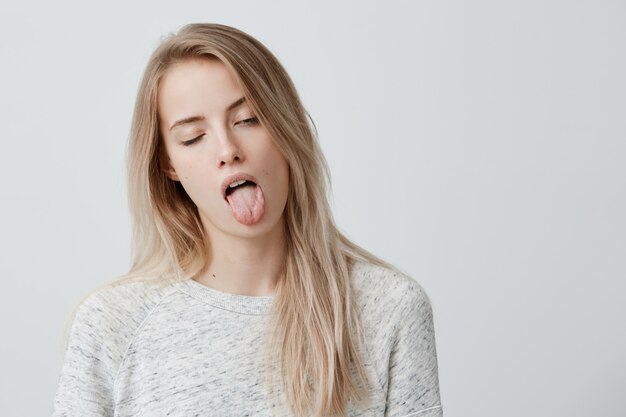 Reacción humana negativa, sentimientos y actitud. Retrato de mujer rubia molesta y aburrida en ropa casual haciendo muecas, sacando la lengua, parpadeando, sintiendo náuseas debido al mal olor