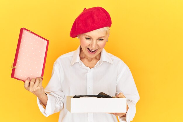 Reacciones y emociones humanas positivas. Mujer de mediana edad emocional de moda con gorro rojo que exclama con entusiasmo, con una mirada de asombro y alegría, abriendo la caja con un regalo inesperado, manteniendo la boca abierta