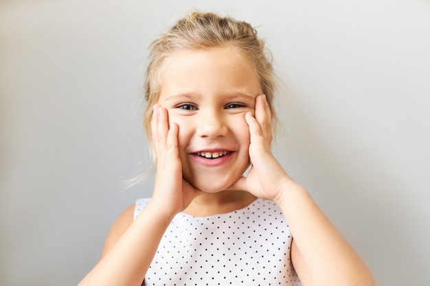 Reacciones y sentimientos humanos genuinos. Retrato de hermoso niño lindo en vestido punteado manteniendo las manos en sus mejillas regordetas, habiendo feliz expresión facial feliz, emocionado con el regalo de cumpleaños