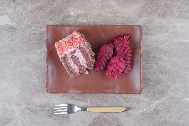 Una rebanada de pastel y conos de pino rojo en un plato junto a un tenedor en mármol