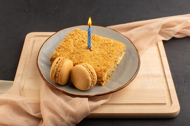 Una rebanada de pastel de vista frontal delicioso y horneado dentro de la placa con vela. Macarons en el escritorio de madera y pastel oscuro galleta dulce de azúcar