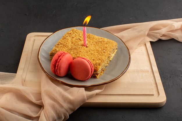 Una rebanada de pastel de vista frontal delicioso y horneado dentro de la placa con velas y macarons en el escritorio de madera y azúcar de galleta de pastel oscuro
