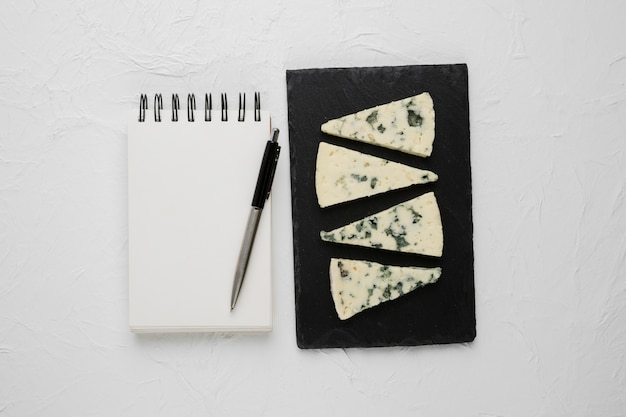 Rebanada de queso azul dispuesta en pizarra negra con cuaderno de espiral vacío y pluma sobre superficie de concreto