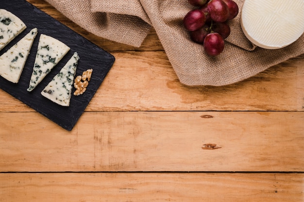 Foto gratuita rebanada de queso gorgonzola; nogal en piedra negra con uvas y queso manchego español sobre textura de arpillera