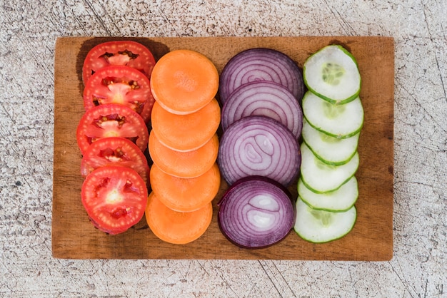 Rebanada de tomates; zanahorias; Cebolla y pepino dispuestos en tablero de madera sobre el fondo de hormigón