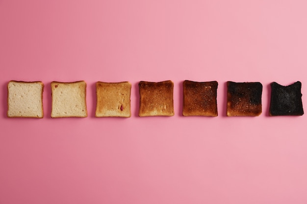 Rebanadas de pan en diferentes etapas de tostado. Rodajas tostadas crujientes dispuestas en una fila sobre fondo rosa. El último en completamente quemado. Haciendo tostadas. De tostado a carbonizado. Vista de arriba hacia abajo