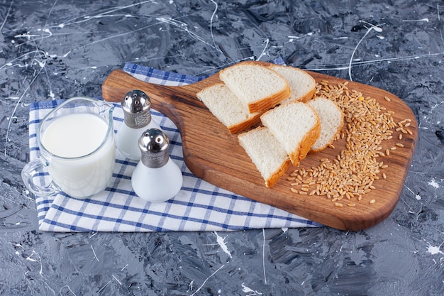 Rebanadas de pan y grano en la tabla de cortar junto a la sal y la leche en una toalla, sobre la mesa azul.
